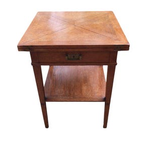 Op de afbeelding: Een kleine, vierkante houten bijzettafel met een lade en een onderste plank. De tafel is gemaakt van lichtbruin hout met een geometrisch patroon op het tafelblad. Hij heeft vier slanke poten en een kleine messing ladeknop.