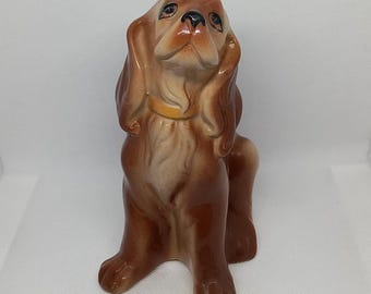 Peut inclure: Une figurine en céramique brune d'un chien aux oreilles tombantes, assis la tête levée. Le chien porte un collier jaune.