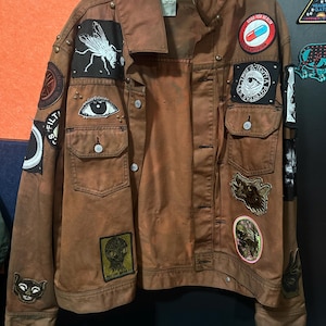 Puede incluir: Chaqueta vaquera marrón adornada con varios parches, incluyendo una mosca, un ojo y una pastilla con la inscripción "GOOD FOR BRAIN". La chaqueta tiene múltiples bolsillos y tachuelas metálicas. La chaqueta está colgada en una percha.