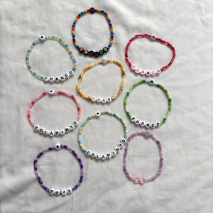 Puede incluir: Una colección de pulseras de cuentas de colores, con nombres e iniciales deletreados con cuentas de letras blancas. Las pulseras son en tonos de rosa, azul, verde, amarillo, morado y arcoíris.