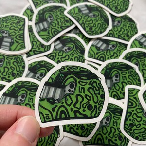 May include: A close-up of a green and black patterned sticker of a racing helmet. The helmet has a black visor and a green and black camouflage pattern.
