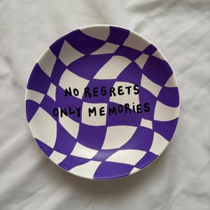 May include: A white ceramic plate with a purple and white checkered pattern. The plate has the words "NO REGRETS ONLY MEMORIES" in black lettering.