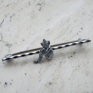 May include: A silver bar brooch with a small, seated figure as its focal point. The pin has a textured bar and a detailed figurine, possibly metal. This vintage-style pin is ideal for adding a touch of character to garments or accessories.