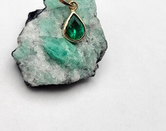 May include: A teardrop-shaped emerald gemstone pendant set in gold. The pendant is resting on a piece of green and white rock.