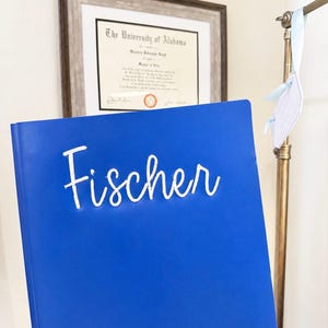 May include: A bright blue binder featuring the name Fischer in white cursive lettering on the front cover. The binder has a smooth, solid-colored surface and a vertical spine on the left side, presenting a clean and simple design for document storage.