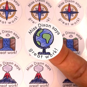 Puede incluir: Una colección de pegatinas redondas con varios diseños y el texto "Miss Dixon says great work!". Los diseños incluyen una brújula, un globo terráqueo, una cascada y un volcán. Las pegatinas son blancas con ilustraciones coloridas.