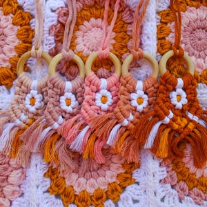 May include: Five macrame necklaces with wooden rings and daisy-like designs in shades of peach, orange, and white. The necklaces have fringe and are displayed on a crocheted blanket with a floral pattern.