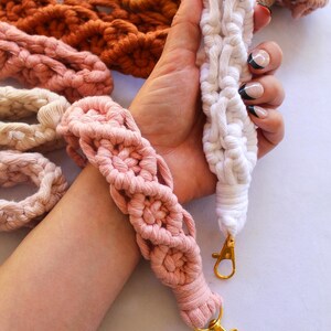May include: Macrame wristlets in various colors, including white, pink, and orange. The wristlets have a woven design and gold-colored clasps. Other macrame items are visible in the background.