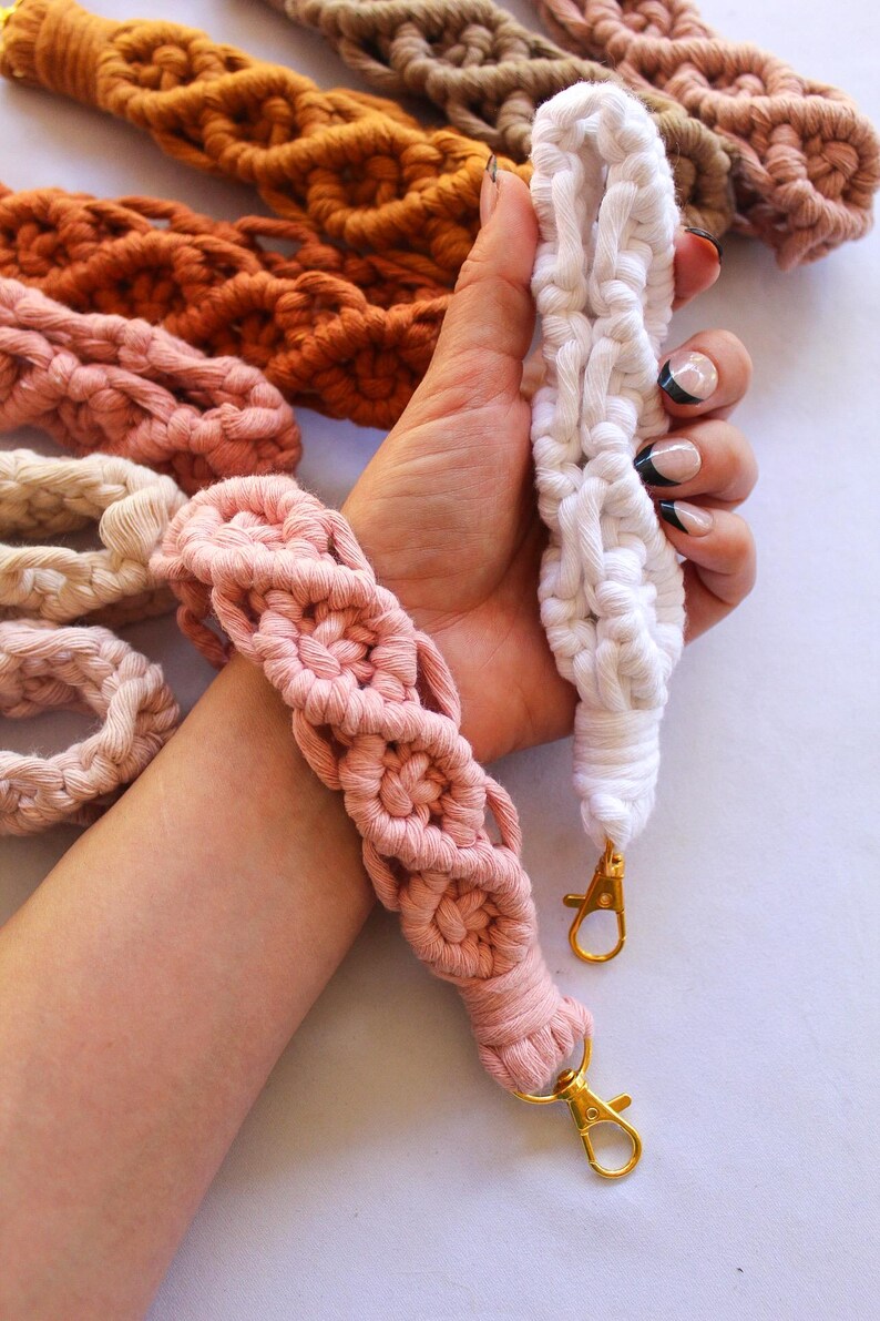 May include: Macrame wristlets in various colors, including white, pink, and orange. The wristlets have a woven design and gold-colored clasps. Other macrame items are visible in the background.