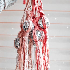 May include: A cluster of macrame plant hangers in shades of pink and white, each holding a small disco ball. The hangers are knotted and feature long, fringed tassels. The disco balls are made of mirrored tiles.