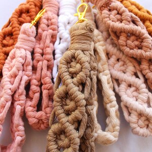 May include: A collection of handmade macrame wristlets in various colors, including shades of brown, pink, and white. Each wristlet features a woven design and a gold-colored clasp for attaching keys or other small items.