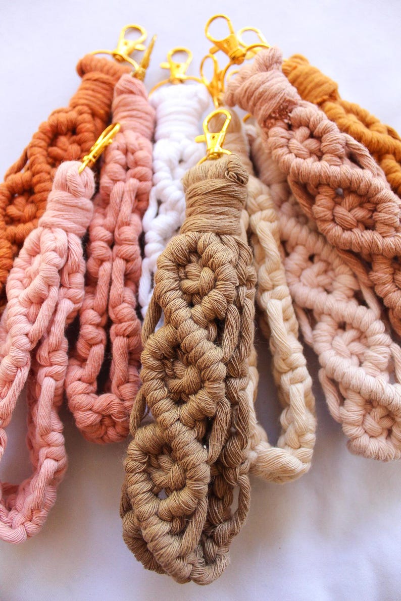 May include: A collection of handmade macrame wristlets in various colors, including shades of brown, pink, and white. Each wristlet features a woven design and a gold-colored clasp for attaching keys or other small items.