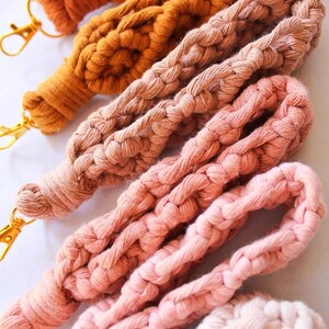 May include: Close-up of several macrame lanyards in various shades of brown, orange, and pink. Each lanyard is made of thick, braided cotton cord and features a gold-colored clasp. The lanyards are arranged diagonally, showcasing their texture and color variations.