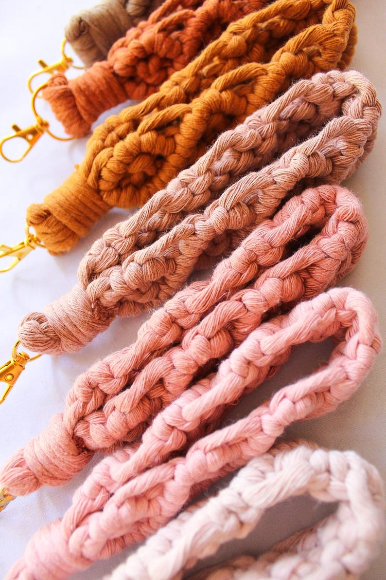 May include: Close-up of several macrame lanyards in various shades of brown, orange, and pink. Each lanyard is made of thick, braided cotton cord and features a gold-colored clasp. The lanyards are arranged diagonally, showcasing their texture and color variations.