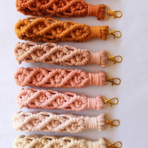 May include: A collection of macrame keychains in various colors, including beige, orange, and pink. Each keychain features a woven design and a gold-colored clasp for attaching keys or other items.