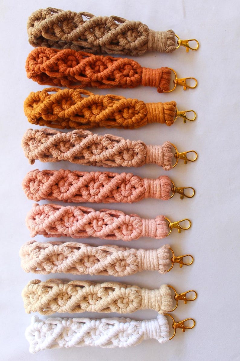 May include: A collection of macrame keychains in various colors, including beige, orange, and pink. Each keychain features a woven design and a gold-colored clasp for attaching keys or other items.