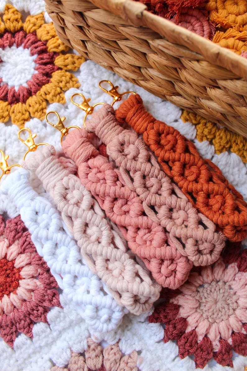 May include: Close-up of five macrame keychains in shades of white, pink, and orange, each with a gold-colored clasp. The keychains are arranged on a colorful, crocheted blanket. A woven basket is in the background.