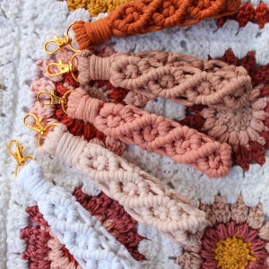 May include: A collection of macrame keychains in various colors, including white, peach, and rust, with gold-colored clasps. The keychains are displayed on a crocheted blanket with a floral pattern in shades of white, peach, and mustard.