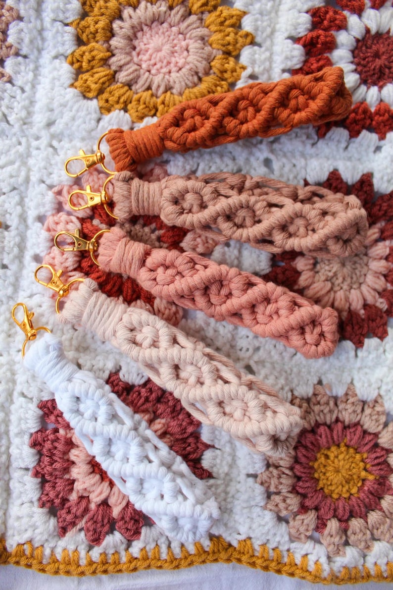 May include: A collection of macrame keychains in various colors, including white, peach, and rust, with gold-colored clasps. The keychains are displayed on a crocheted blanket with a floral pattern in shades of white, peach, and mustard.