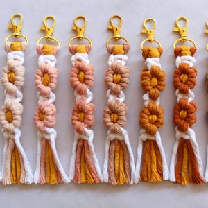 May include: A collection of eight macrame keychains with gold-colored clasps. Each keychain features a flower design in shades of white, peach, orange, and brown. The tassels at the bottom have matching color schemes.