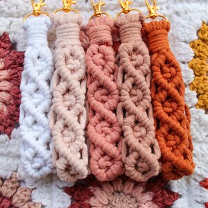 May include: Five macrame keychains in shades of white, peach, pink, and orange, each with a gold-colored clasp. The keychains are arranged on a crocheted blanket with a floral pattern in similar colors.