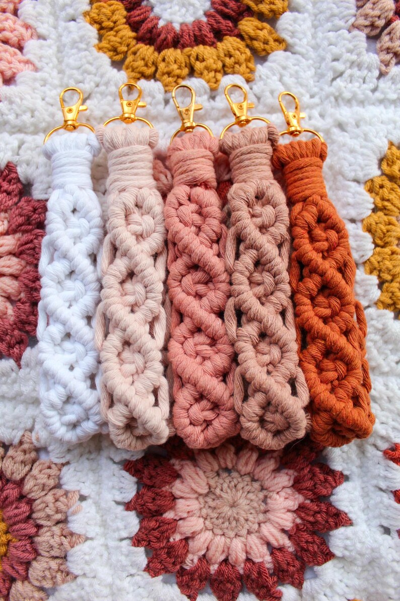 May include: Five macrame keychains in shades of white, peach, pink, and orange, each with a gold-colored clasp. The keychains are arranged on a crocheted blanket with a floral pattern in similar colors.
