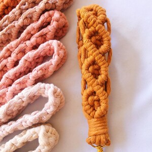 May include: Close-up of a macrame lanyard in a warm orange hue, featuring a series of woven flower-like patterns. Several other macrame cords in shades of brown, peach, and cream are arranged nearby. The lanyard has a gold-colored clasp.