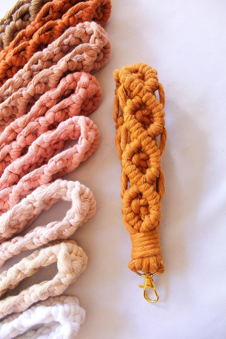 May include: Close-up of a macrame lanyard in a warm orange hue, featuring a series of woven flower-like patterns. Several other macrame cords in shades of brown, peach, and cream are arranged nearby. The lanyard has a gold-colored clasp.