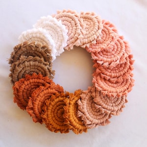 May include: A circular arrangement of handmade macrame coasters in a gradient of colors, from white to peach, orange, and brown. Each coaster features a spiral design with fringed edges, creating a textured and decorative look. The coasters are arranged in a circle.
