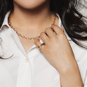 May include: Gold-toned jewelry set featuring a necklace, bracelet, and ring. The necklace and bracelet have an oval link design with embedded clear stones. The ring has a spiral design. The jewelry is worn over a white collared shirt.