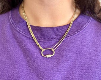 Puede incluir: Un collar de cadena de oro con un pequeño colgante de forma ovalada que presenta una fila de piedras transparentes. El collar está superpuesto a una segunda cadena de oro más fina.