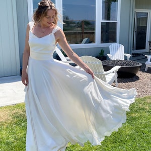 May include: A white sleeveless wedding dress with a sweetheart neckline and a flowing skirt. The dress is being worn by a person outdoors in front of a house.