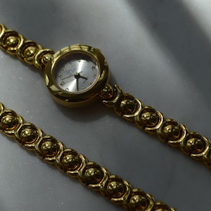 May include: A gold-toned wristwatch with a silver face and matching gold band. The band features a repeating pattern of connected, spherical links. The watch is displayed on a light-colored surface, with soft lighting.