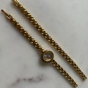 May include: A gold-tone watch with a round face and a linked bracelet. The watch is on a white surface.