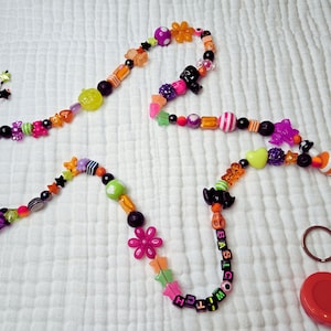 May include: A beaded lanyard with a Halloween theme. The lanyard features a variety of colorful beads, including stars, flowers, and pumpkins. The text "I'M BASIC, WITCH" is spelled out with letter beads. A black clasp and a red retractable badge holder are also included.