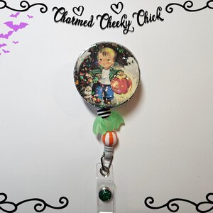 May include: A Halloween themed retractable badge holder with a green and white pumpkin charm, a white and orange striped bead, and a round image of a boy holding a pumpkin. The image is surrounded by glitter and the text "Charmed Cheeky Chick" is at the top of the image.