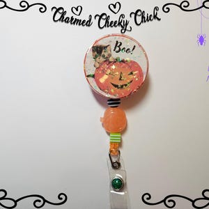 May include: A Halloween themed retractable badge holder featuring a pumpkin and kitten design. The pumpkin is orange with a glittery finish and says "Boo!" The kitten is black and white and is peeking out from behind the pumpkin. The badge holder has a clear plastic strap and a green glitter button.