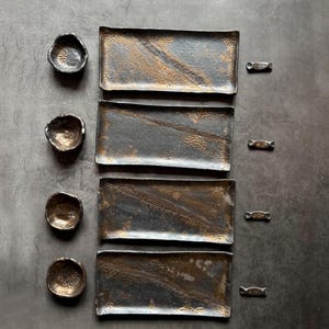 May include: A set of handmade ceramic serving dishes. The set includes four rectangular plates with a textured surface and a dark gray and gold finish, along with four small, round bowls and four small, candy-shaped dishes.