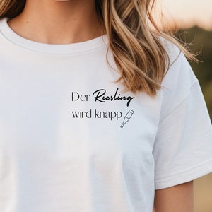 Könnte beinhalten: Weißes T-Shirt mit dem Text "Der Riesling wird knapp" und einem kleinen Bild einer Weinflasche.