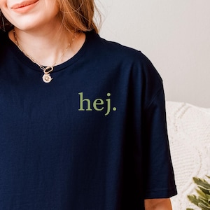 Pode incluir: Camiseta azul marinho com a palavra "hej." em verde no lado direito do peito. A camiseta é de estilo gola redonda clássica e é feita de um material macio. A modelo está usando jeans azul claro.