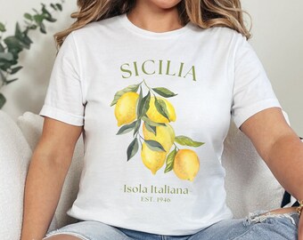 May include: White t-shirt with a watercolor illustration of lemons and the text "SICILIA" and "Isola Italiana EST. 1946". The lemons are yellow with green leaves. The shirt is worn with blue jeans.