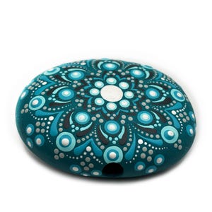 Puede incluir: Una piedra pintada a mano de color turquesa con un diseño de mandala. El mandala presenta un círculo blanco central rodeado de patrones turquesa, azul claro y gris. La piedra es de forma ovalada y tiene una superficie lisa.