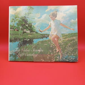 Puede incluir: Un catálogo de regalos vintage de Top Value Stamps con una niña con un vestido blanco saltando sobre un arroyo en un campo de flores. La portada del catálogo tiene un cielo azul con nubes blancas y un campo verde con un granero rojo en el fondo.