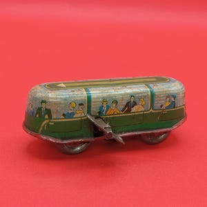 May include: A vintage tin toy bus featuring a green and blue exterior with illustrations of passengers visible through painted windows. The toy has a metal winding key on the side and small wheels, with a yellow arrow graphic on the roof.