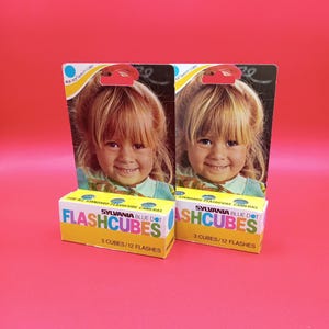 May include: Two packs of Sylvania Blue Dot Flashcubes, each containing 3 cubes for a total of 12 flashes. The packaging features a smiling young girl with blonde hair and a blue dot logo.