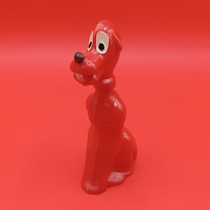 Könnte beinhalten: Eine rote Cartoon-Hundefigur mit weißen Augen und einer schwarzen Nase. Der Hund sitzt aufrecht, der Kopf leicht zur Seite geneigt. Der Hintergrund ist einfarbig rot.