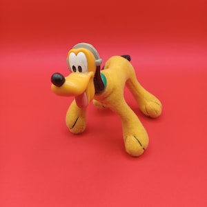Könnte beinhalten: Ein gelbes Plüschtier von Pluto dem Hund, das ein graues Stirnband mit grünem Akzent trägt. Das Spielzeug steht auf einem roten Hintergrund.