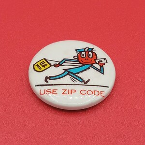 Op de afbeelding: Een ronde witte button met een cartoonfiguur met een rood gezicht en blauw uniform die rent terwijl deze een gele U.S. Mail-tas en een brief draagt. Onder het figuur staat de tekst USE ZIP CODE in rode hoofdletters afgedrukt.