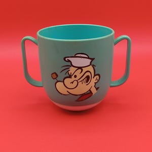 May include: A turquoise blue plastic cup with two handles. The cup features a cartoon image of Popeye the Sailor Man with his signature pipe and a surprised expression.