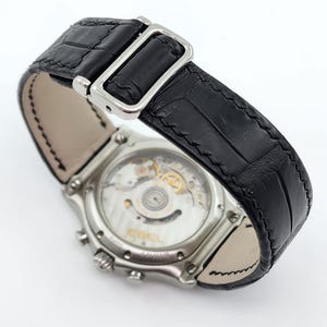 May include: A silver-toned wristwatch with a black leather band. The watch features a visible mechanism on the back and a black, textured band with a silver buckle. The watch is set against a white background.